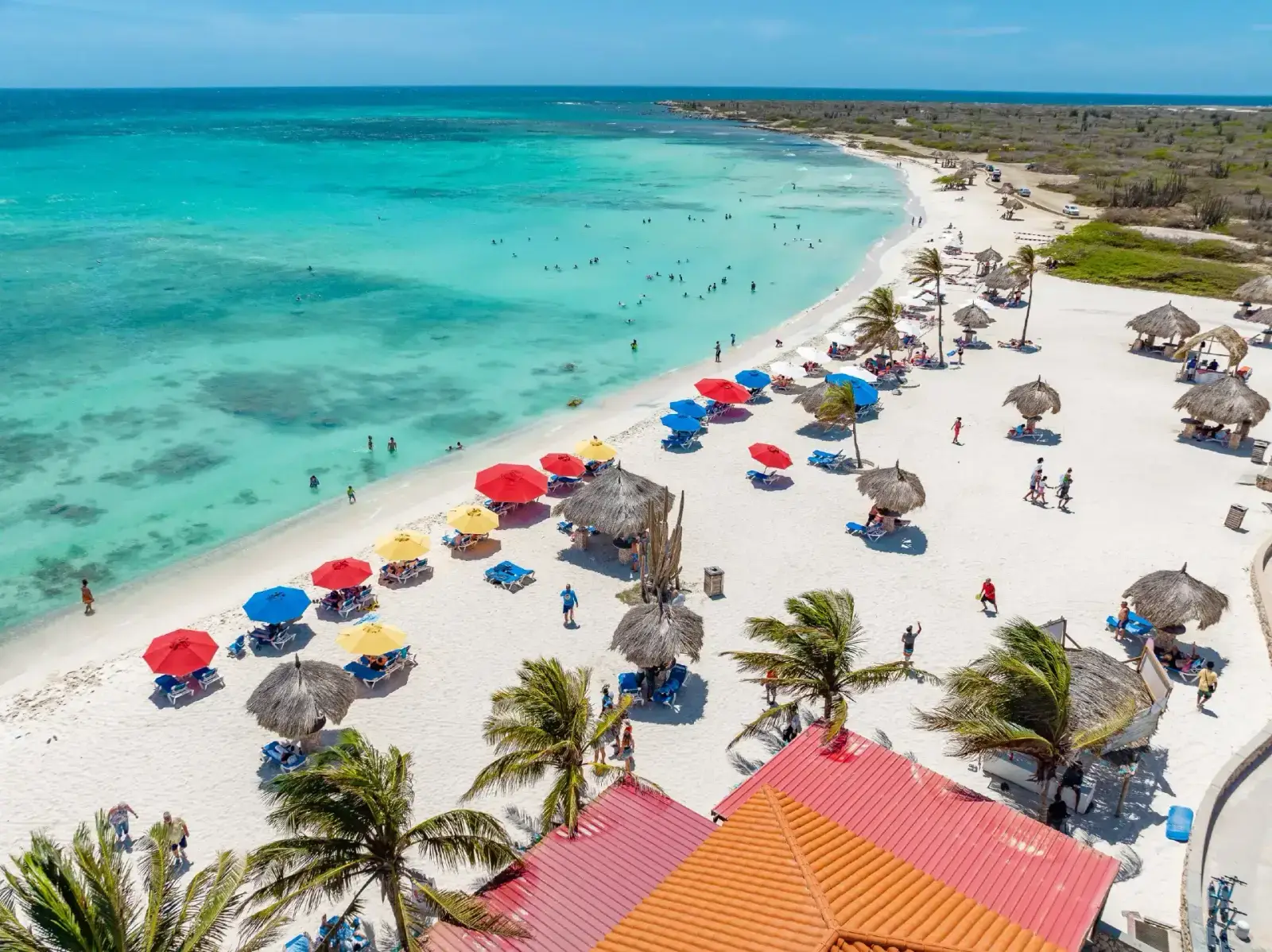 Aerial view of a busy white-sand Aruba beach with turquoise water and colorful umbrellas